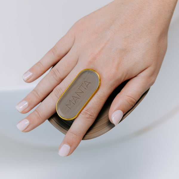 A close-up of a hand with pale pink manicured nails resting on a round wooden bath brush, with a gold-toned oval metal ring on the back of the hand that reads 'MANTA'.
