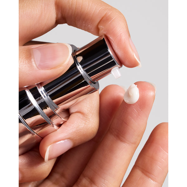 A close-up of a hand pressing the pump of a metallic cosmetic bottle, with a small dollop of white cream visible on another finger against a light gray background.
