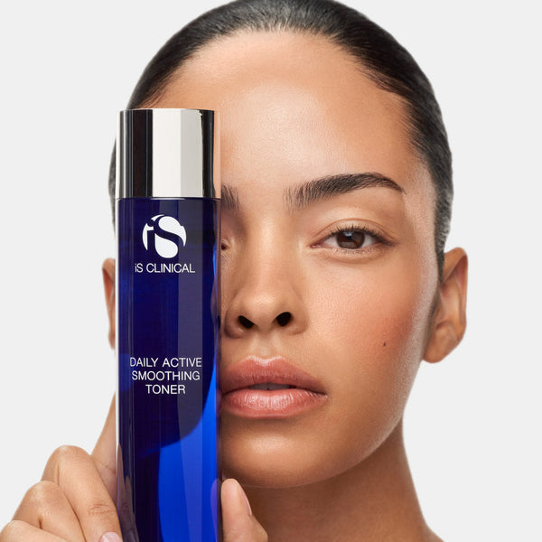 A woman holds a bottle of IS Clinical Daily Active Smoothing Toner next to her face against a light gray background.
