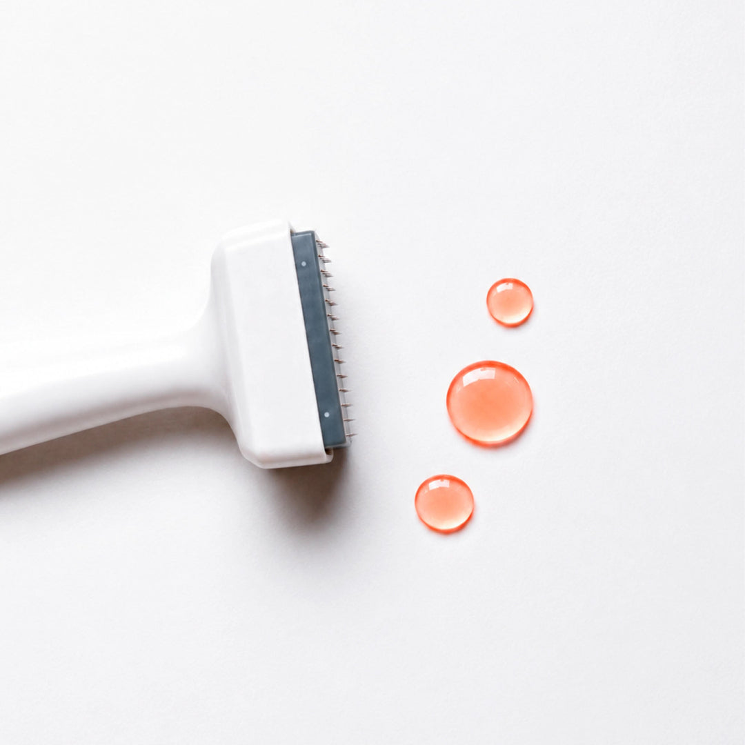 A close-up of a white pet grooming brush with a dark gray metal edge, positioned next to three clear droplets of orange liquid on a clean white background.