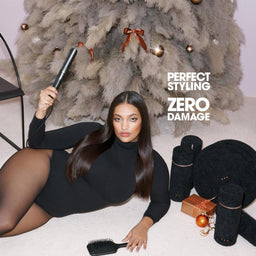 A woman in a black dress and sheer black tights reclines on a light floor in a styled room, holding a black hair tool. A large gray textured tree, black chair, gold box, and several black hair accessories are arranged around her.