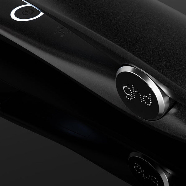 A close-up of a black ghd hair straightener with its power cord, showing the brand logo on the control dial against a dark reflective background.