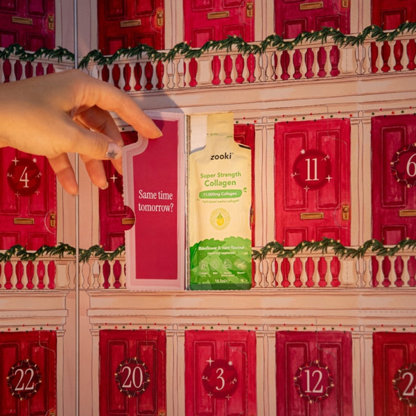 A hand opens a window on a decorative advent calendar with numbered doors, revealing a carton of zooki Super Strength Collagen.