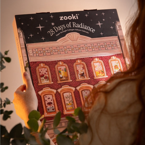 A person holds a Zooki 28 Days of Radiance luxury chocolate advent calendar box. The box features a red brick building design with illustrated windows and a black starry top banner.