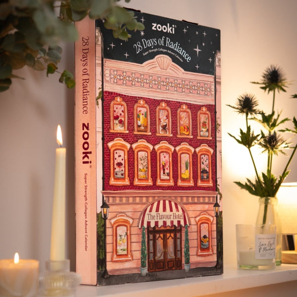 A hardcover book titled 'zooki 28 Days of Radiance' stands upright on a shelf, surrounded by candles and a glass vase with flowers in a warm, styled interior setting.
