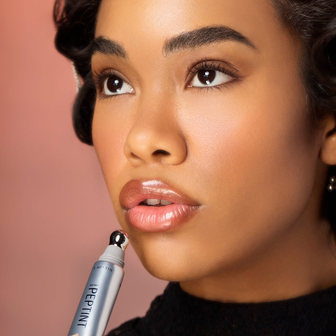 A close-up of a woman with vintage-style curled hair holding a silver cosmetic tube near her lips against a soft pink background.