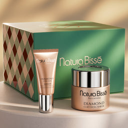 Nature Bisse Diamond Cocoa Shot skincare set featuring a green gift box, a peach-colored cream tube, and a matching jar with a silver lid, displayed on a white pedestal against a warm beige background with soft leaf shadows.