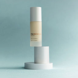 A bottle of Omorovicza Body Vitamin B5 & Ceramide moisturizer stands upright on a white cylindrical pedestal against a soft light blue background.