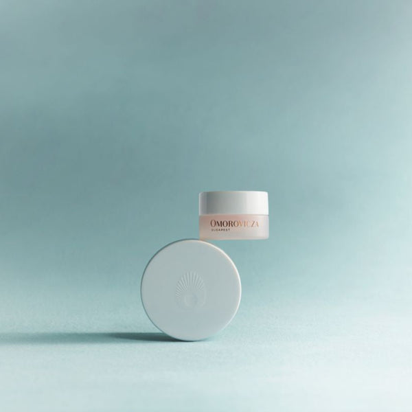 A small cosmetic jar with a white lid floating above a matching round white container on a soft blue gradient background. The jar label shows the brand name UNIBOYA.