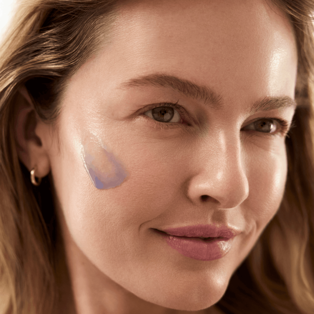 Close-up of a woman's face with a translucent blue-purple cosmetic swatch applied to her cheek, showing a glossy, dewy finish.