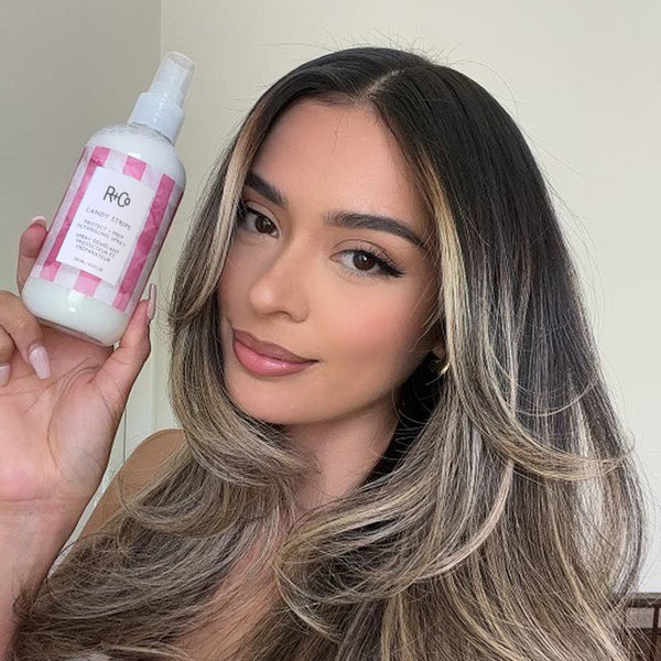 Woman with long, wavy hair holding a bottle of R+Co hair product.