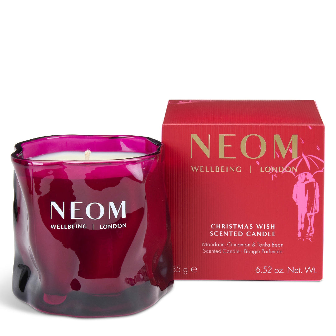 A NEOM Wellbeing London 'Christmas Wish' scented candle in a deep magenta glass jar, shown next to its red packaging box with gold lettering on a white background.