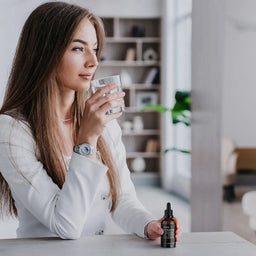 Woman holding a glass of water with a dropper bottle on a table in a modern living room.
