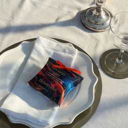 A small packaged favor box labeled 'This Works' and 'Dream Catcher' sits on a white folded napkin atop a white plate with a gold charger, set on a white tablecloth with a glass and candle base nearby.