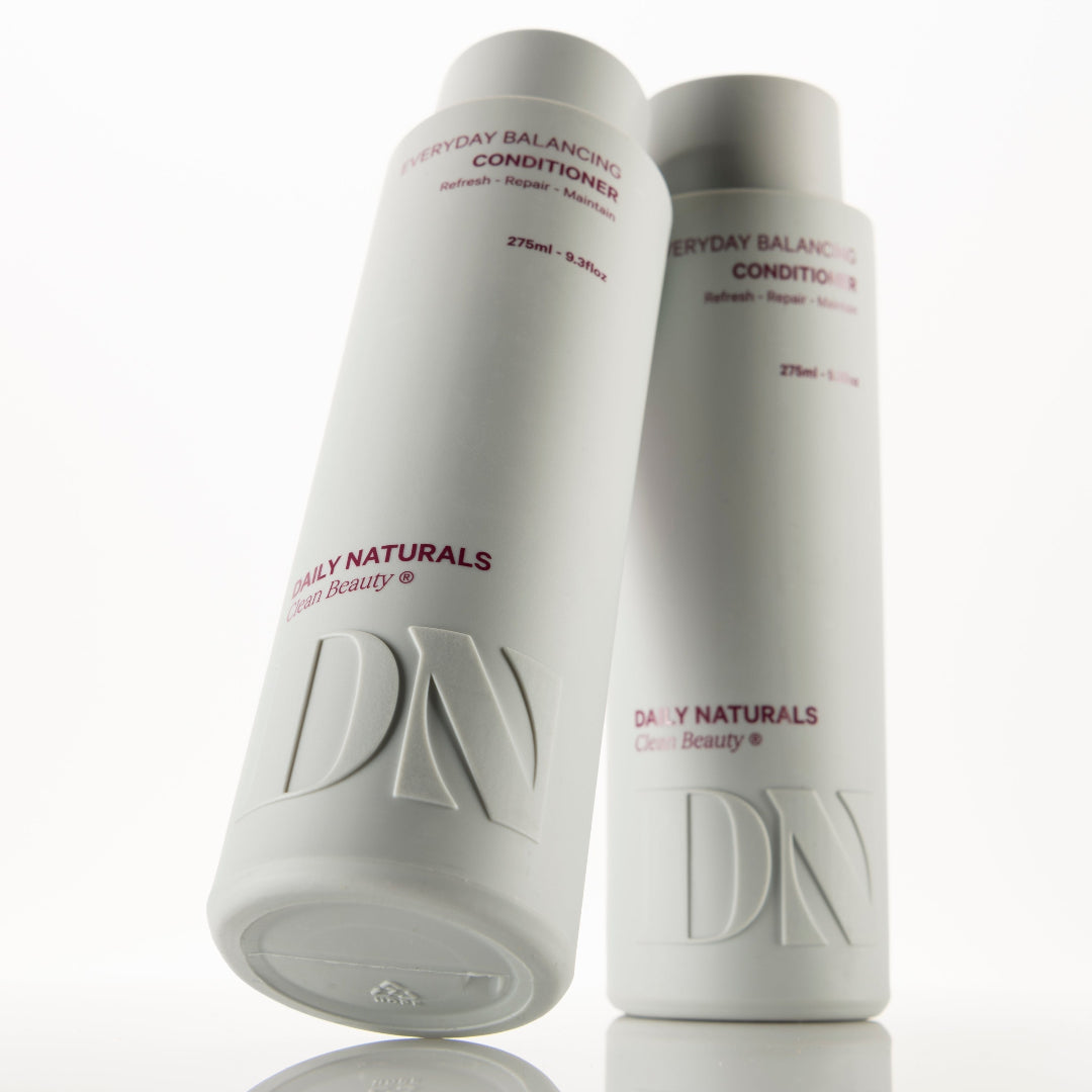 Two white cylindrical bottles of Daily Naturals Everyday Balancing Conditioners on a reflective white surface, with one bottle standing upright and the other tilted in front showing the embossed DN logo.