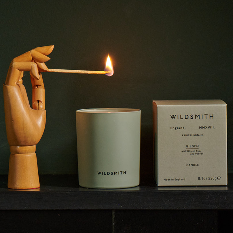 Wooden hand holding a match to light a Wildsmith candle and packaging on a dark background