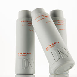 Three white cylindrical bottles of Daily Naturals haircare products, including Intense Hydration Shampoo and Intense Hydration Conditioner, arranged on a reflective white surface.