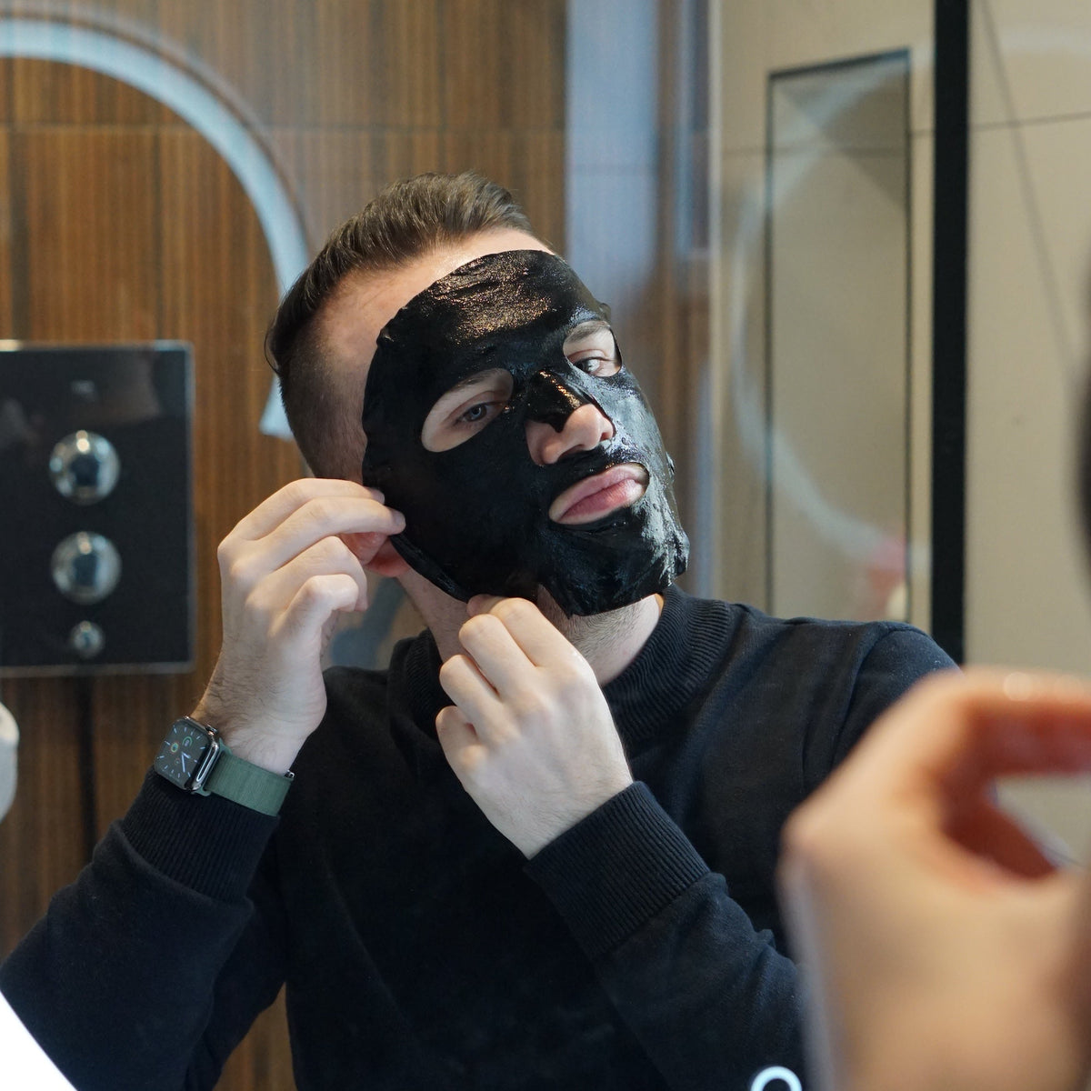 Man applying a black face mask in front of a mirror.