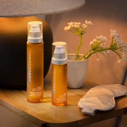 Two bottles of Moroccanoil product on a wooden surface with a plant in the background.