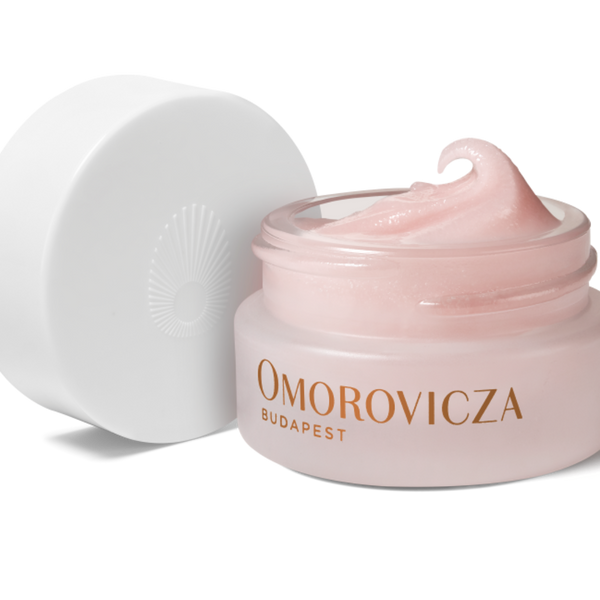 A jar of Omorovicza Budapest cream with its white lid leaning against it on a clean white background. The jar is a soft pale pink, filled with a matching pale pink cream, and features the brand name in gold lettering.