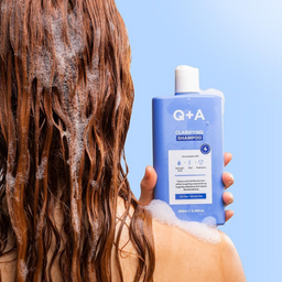 Person with soapy hair holding a bottle of Q+A shampoo against a blue background