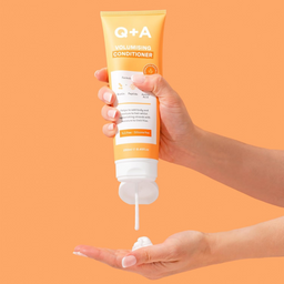 Hand holding a tube of Q+A Volumising Conditioner with a visible pump against an orange background