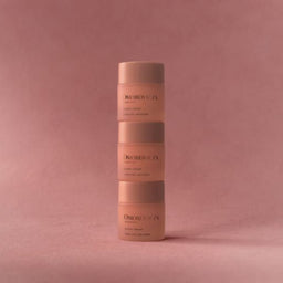 Three stacked cosmetic jars from the brand D'Orbitalia, featuring a soft peachy-pink color with dark brown lettering, set against a matching dusty rose background.
