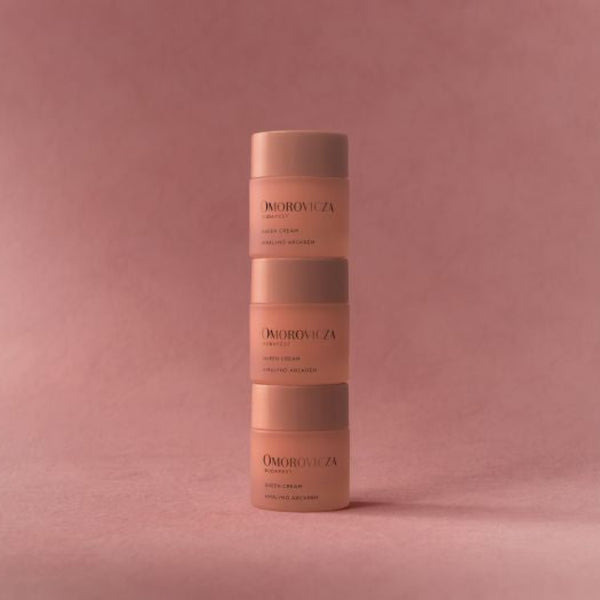 Three stacked cosmetic jars from the brand D'Orbitalia, featuring a soft peachy-pink color with dark brown lettering, set against a matching dusty rose background.