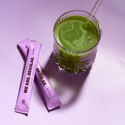 A glass of green drink with a clear straw sits on a light lavender surface next to two pink sachets printed with 'WE ARE. REGULAR.