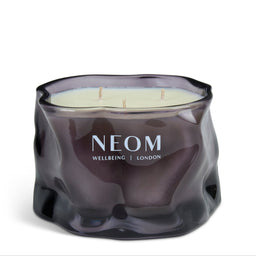 A NEOM Wellbeing London scented candle in a wavy, textured dark purple glass jar with three wicks, set against a plain white background.