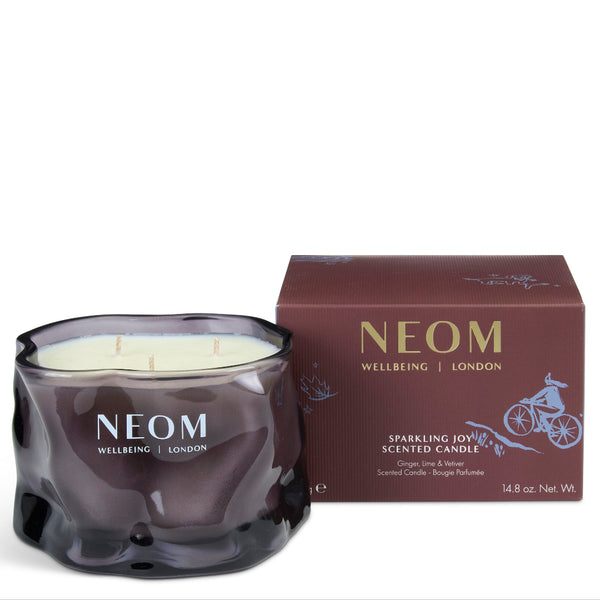 A NEOM Wellbeing London scented candle in a dark, wavy glass jar with cream wax, shown next to its matching dark brown packaging box on a white background.