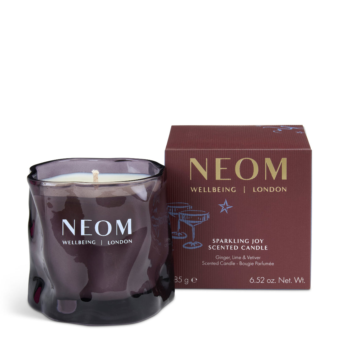 A NEOM Wellbeing London scented candle in a dark purple, textured glass jar next to its dark reddish-brown packaging box, on a clean white background.