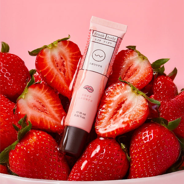 A pink lip tint tube labeled 'fresh body SMOOTH lip tint' standing upright on a pile of fresh sliced strawberries against a solid pink background.