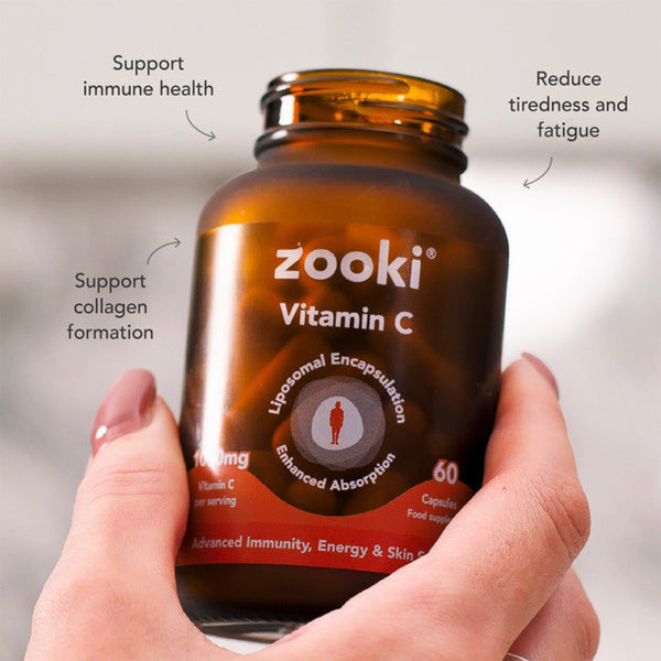 A hand holding an amber brown bottle of zooki Vitamin C capsules, with text callouts around it highlighting benefits like support immune health, reduce tiredness and fatigue, and support collagen formation.