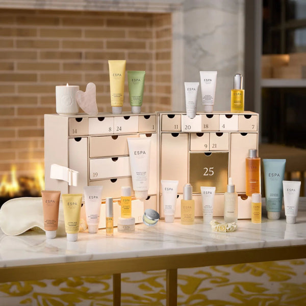 A collection of ESPA skincare and beauty products arranged in front of an open cream-colored advent calendar on a marble-topped table, with a warm fireplace and brick wall in the background.