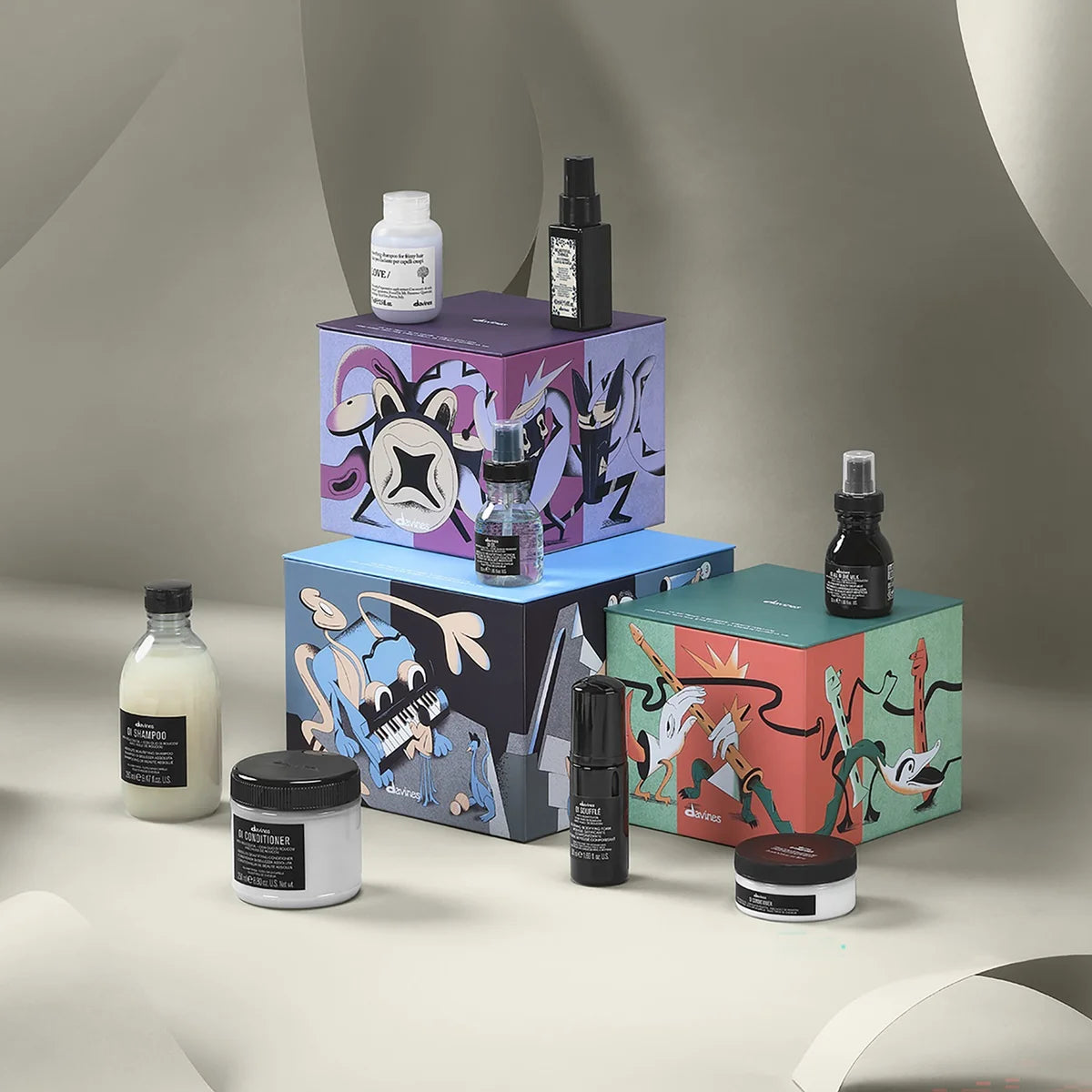 A curated beauty gift set featuring multiple cosmetic products and three colorful illustrated boxes, arranged on a neutral draped studio background. The products include jars, bottles, and a spray bottle with black labels, alongside purple, blue, and green packaging decorated with abstract line-art illustrations.