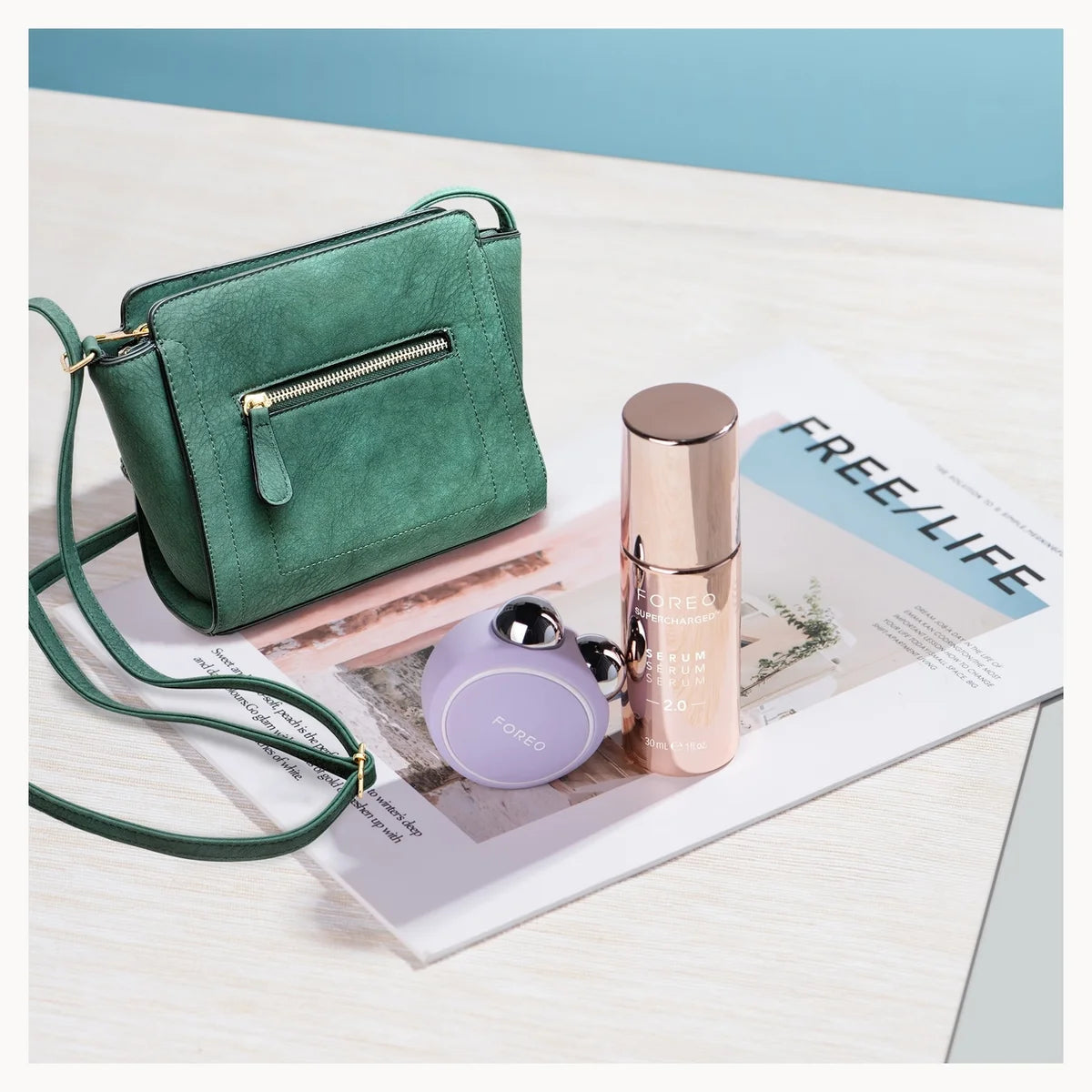 A green suede crossbody handbag with a zippered front pocket and matching strap, styled on a magazine page next to a rose gold cosmetic bottle and a small lavender round pouch.