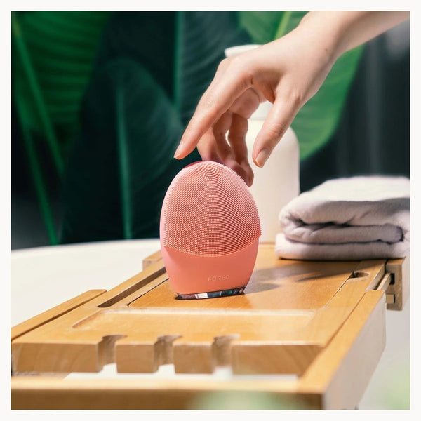 A pink oval facial cleansing brush being touched by a person's hand, resting on a wooden tray with folded towels and a blurred green background.
