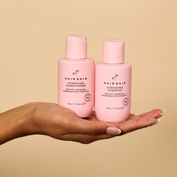 Two pink bottles of Hairgain shampoo and conditioner held in a hand against a beige background